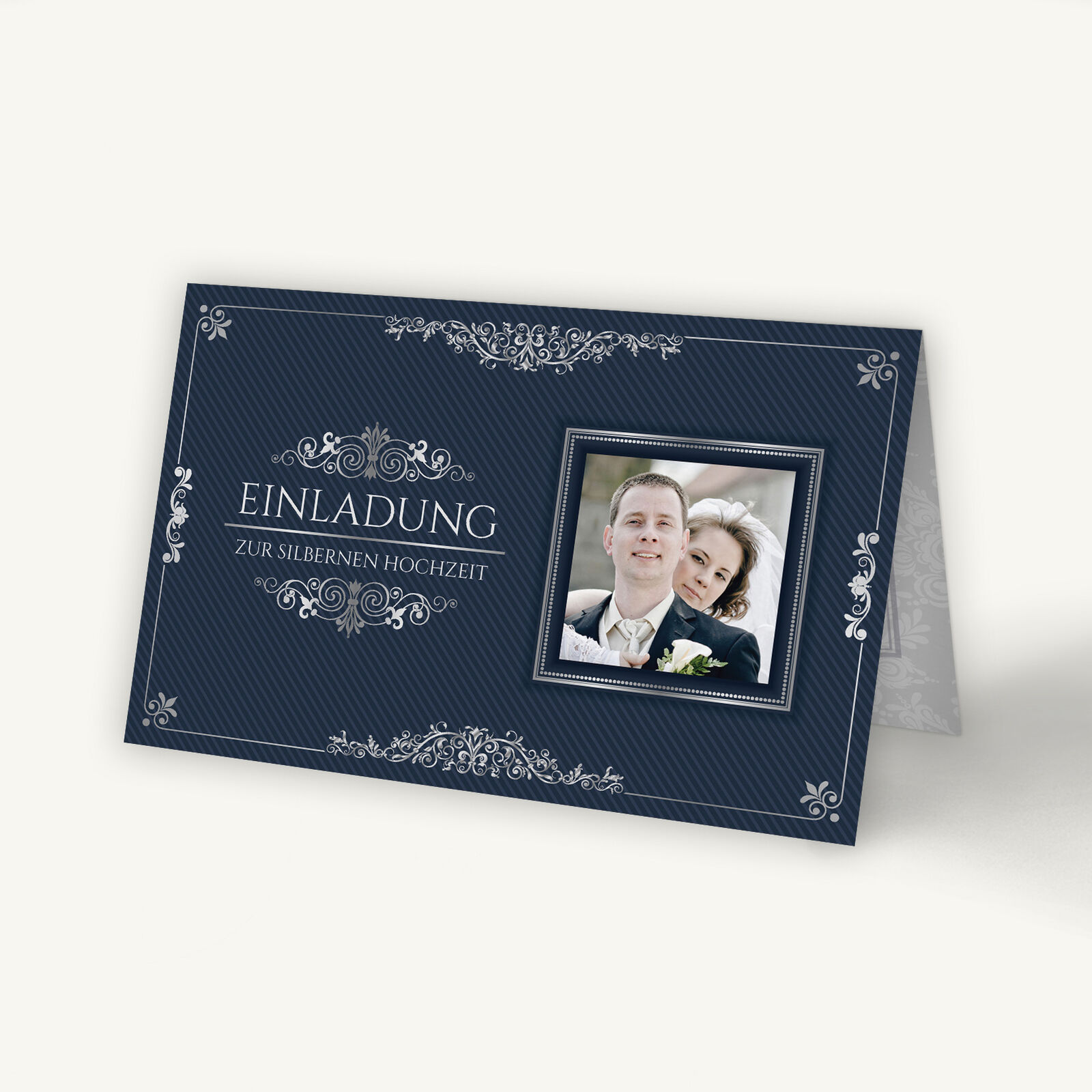Einladungskarte zur Silberhochzeit im eleganten altenglischen Stil. Dunkelblauer Hintergrund mit feinen diagonalen Streifen und silbernen Ornamentverzierungen. In der Mitte steht in klassischer Schrift 'Einladung zur Silbernen Hochzeit'. Rechts befindet sich ein quadratisches Foto eines Paares im Hochzeitsoutfit, eingerahmt von einem silbernen Rahmen.