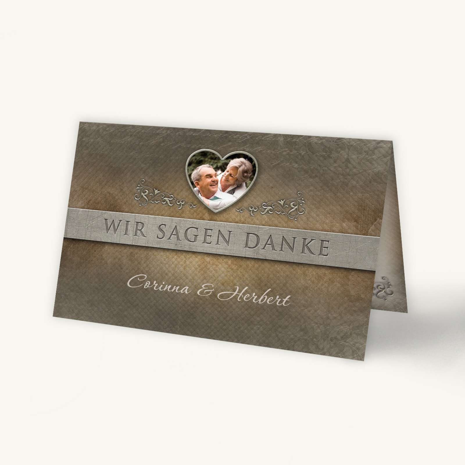 Rustikale Danksagungskarte zur Eisernen Hochzeit im Querformat. Die Klappkarte misst 170 × 110 mm und ist unten offen. Das Design zeigt einen strukturierten, lederähnlichen Hintergrund in warmen Braun- und Grautönen. Mittig verläuft ein breites, silbergraues Band mit feiner Leinenstruktur, auf dem in Großbuchstaben der Schriftzug „WIR SAGEN DANKE“ in eleganter, serifenloser Schrift steht. Über dem Schriftzug befindet sich ein herzförmiger Bildausschnitt mit einem Foto eines älteren Ehepaares, das sich liebevoll anschaut. Feine ornamentale Verzierungen in silbernem Ton rahmen das Herz und den Schriftzug ein. Unter dem Band stehen in geschwungener, weißer Schrift die Namen „Corinna & Herbert“. Das Gesamtbild wirkt edel und würdevoll – passend zum Anlass einer Eisernen Hochzeit.