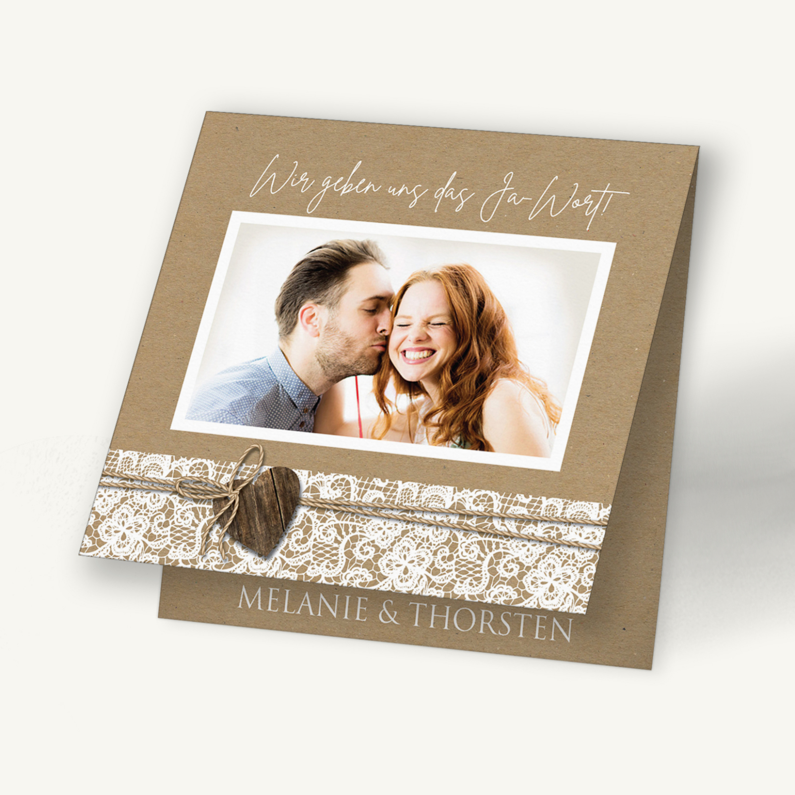 Hochzeitseinladungskarte in natürlichem Kraftpapier-Look mit weißem Spitzenmuster. Oben ein Foto eines lachenden Paares, darunter eine feine Juteschnur mit Herzanhänger aus Holz. Die Namen „Melanie & Thorsten“ runden das Design ab. Der Stil ist rustikal, warm und herzlich – ideal für eine romantische Landhochzeit.