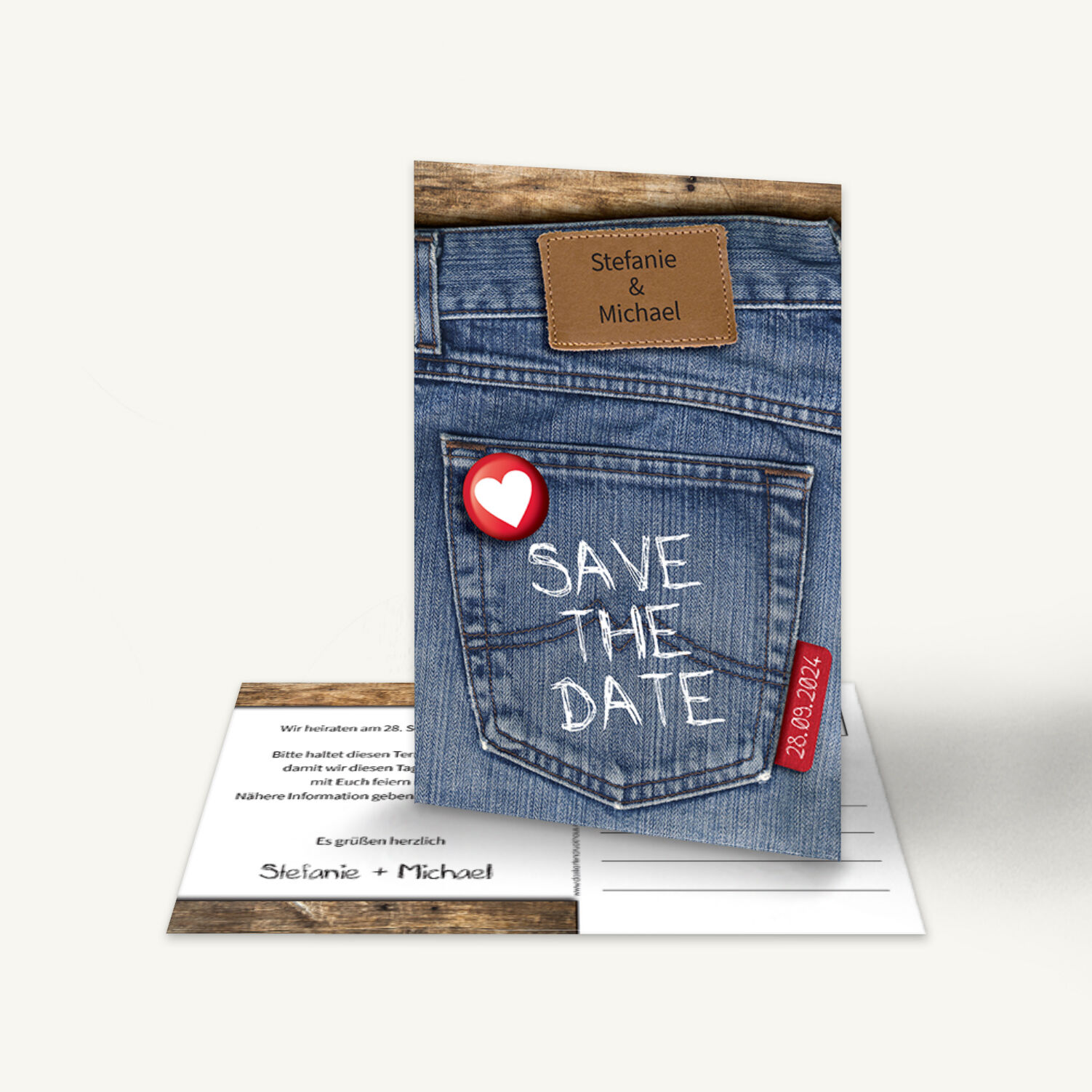 Save-the-Date Karte mit Jeansmotiv und Lederetikett mit den Namen „Stefanie & Michael“. Darunter der Schriftzug „Save the Date“ in Kreideschrift auf der Gesäßtasche, mit einem roten Herz-Button und Datum „28.09.2024“. Lockeres, modernes Design.